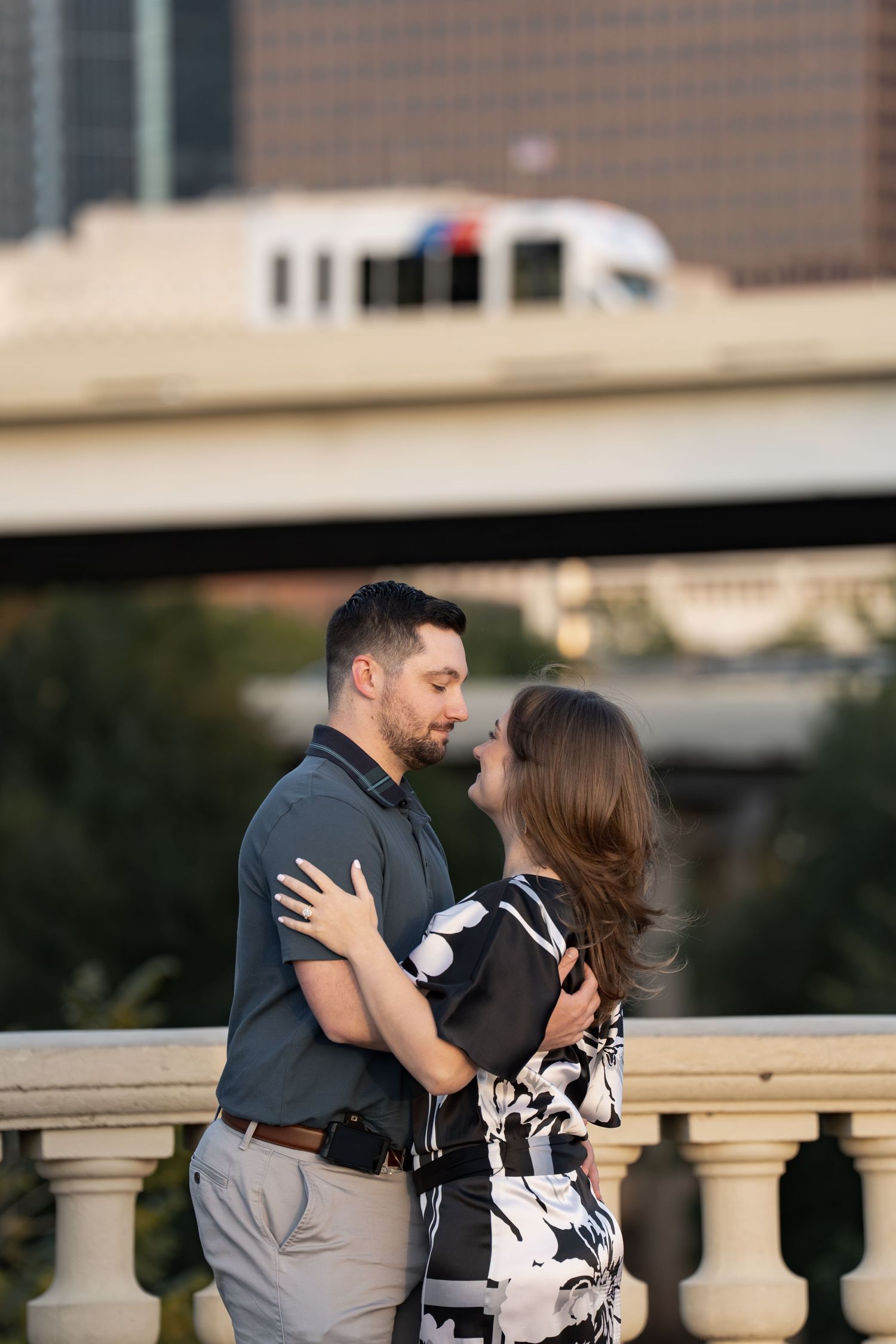 Couple embracing and gazing at each other on a stone balustrade bridge with a city skyline and passing train behind them.