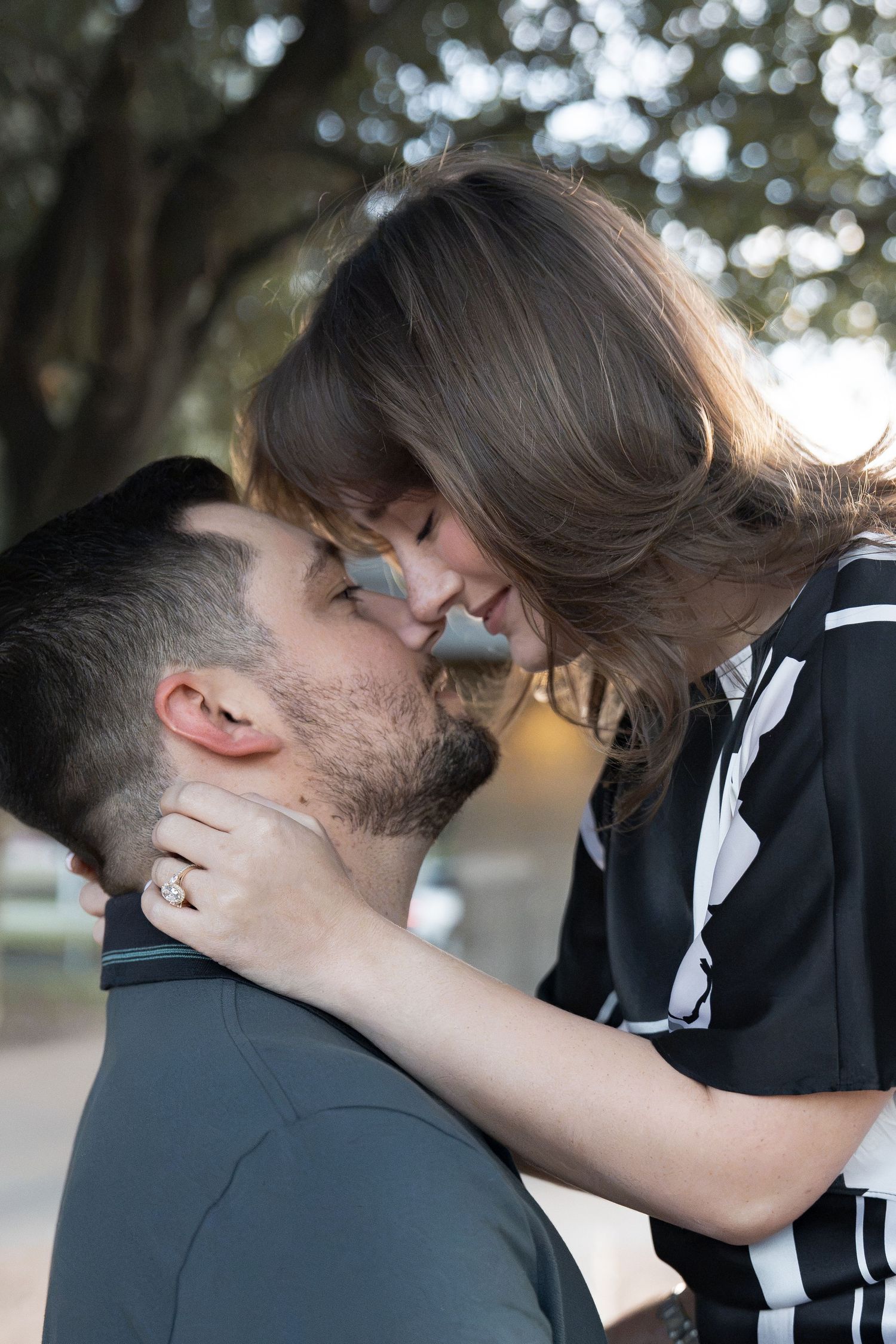 Couple sharing an intimate moment outdoors as a woman with an engagement ring wraps her arm around her fiancé's neck, foreheads touching.