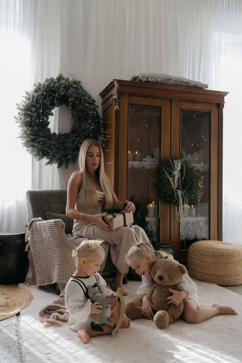 A mother holds a gift while two toddlers play with a teddy bear near a wreath, a wooden cabinet and candles in a cozy Christmas room.