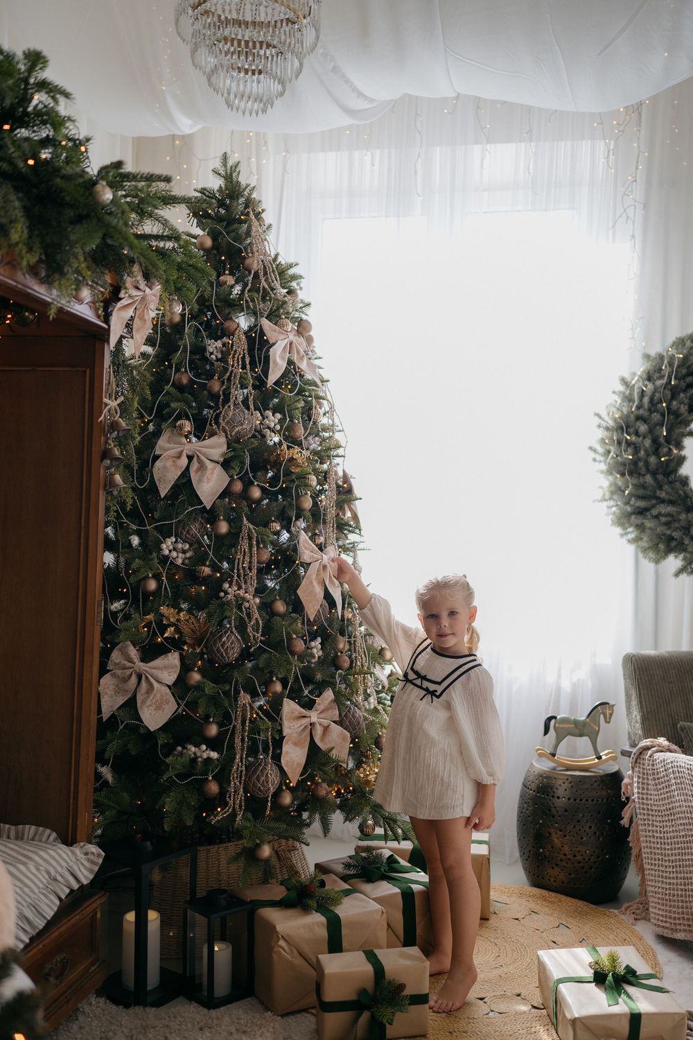 A little girl in a white knit dress reaches toward a Christmas tree trimmed with bows, surrounded by wrapped gifts and a wreath.