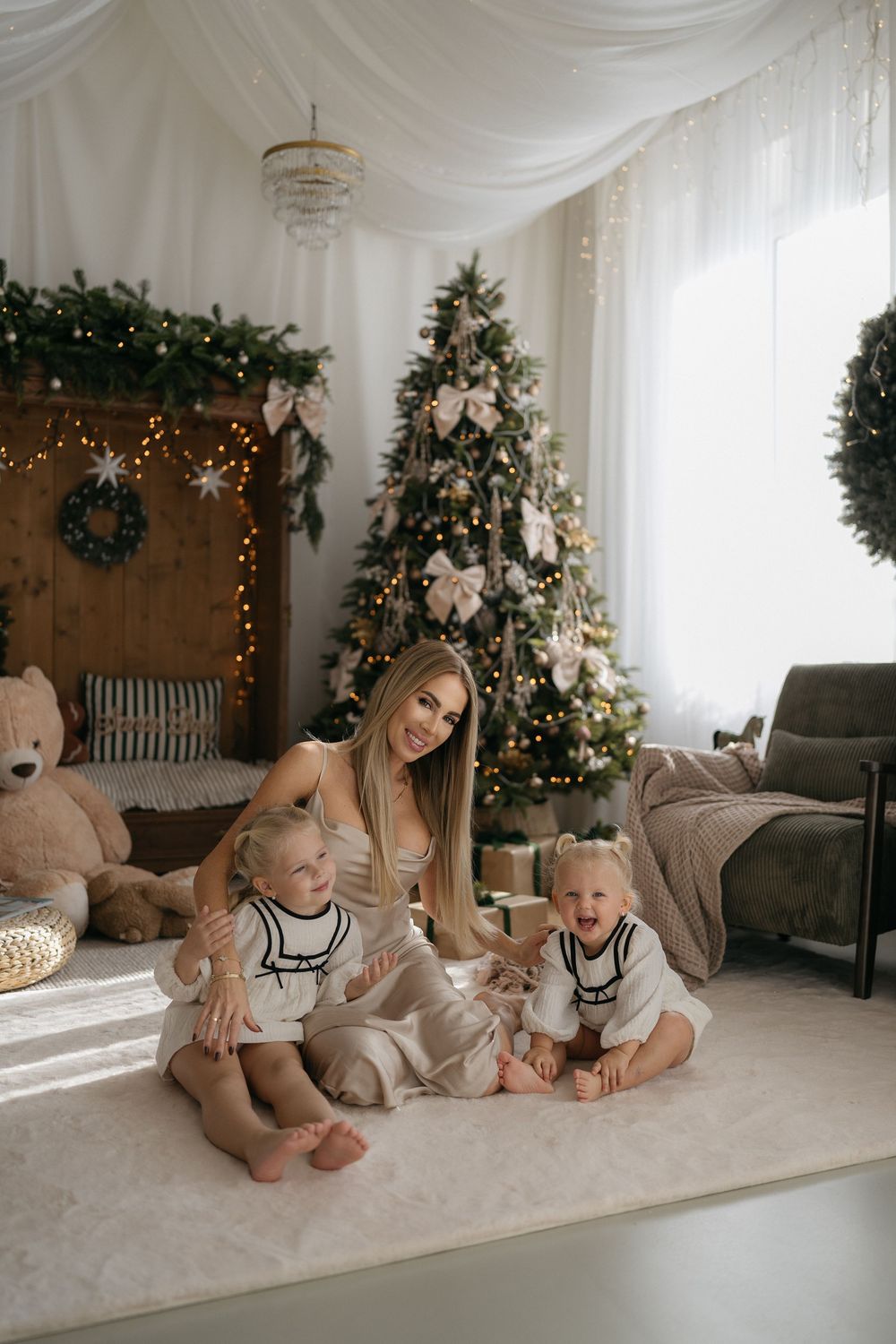 A blonde mother sits on a cream rug hugging two toddlers in white outfits in front of a glowing, ribbon-trimmed Christmas tree.