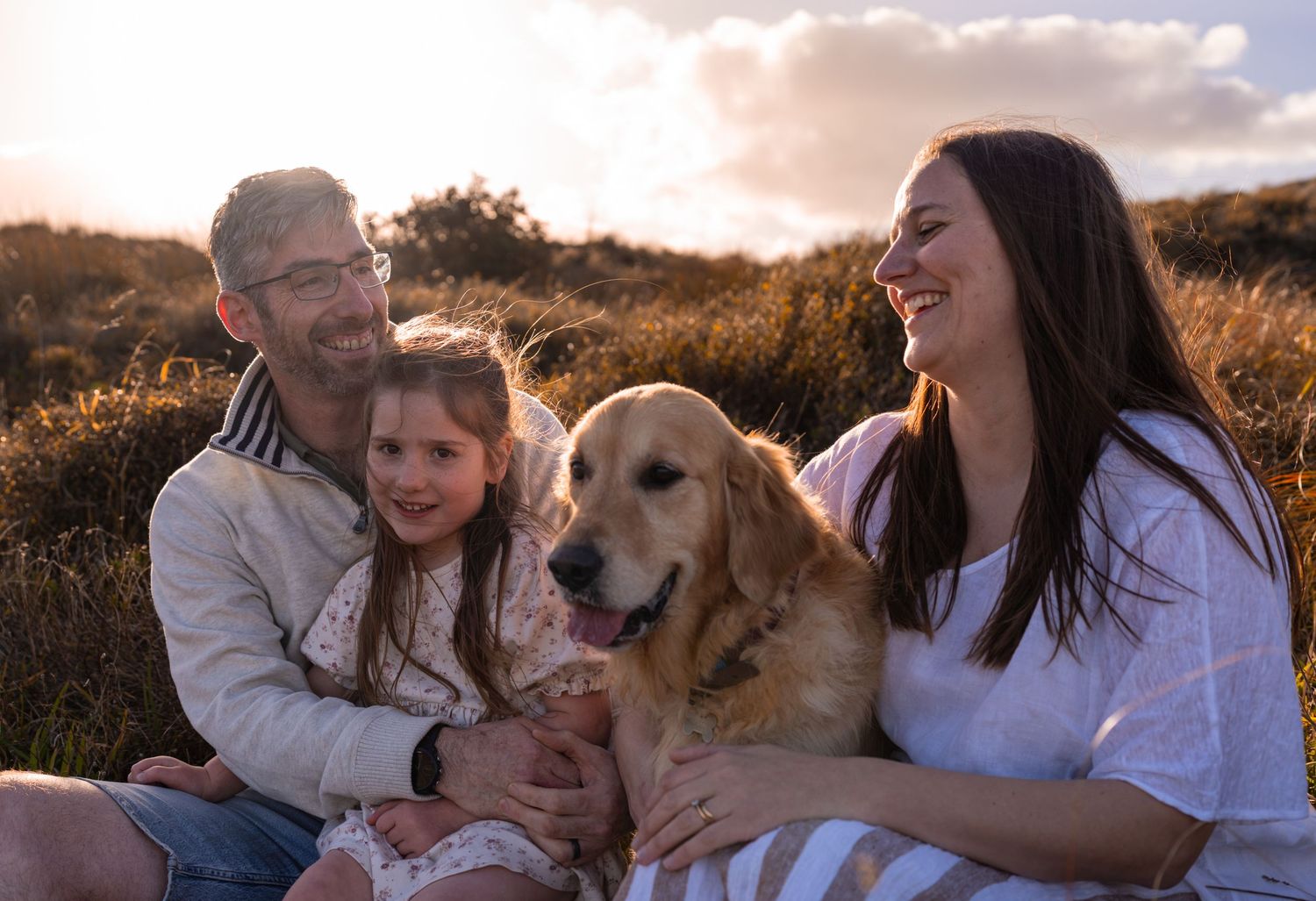 Family with young girl and golden retriever sitting in golden hour sunlight in a grassy field, smiling happily.
