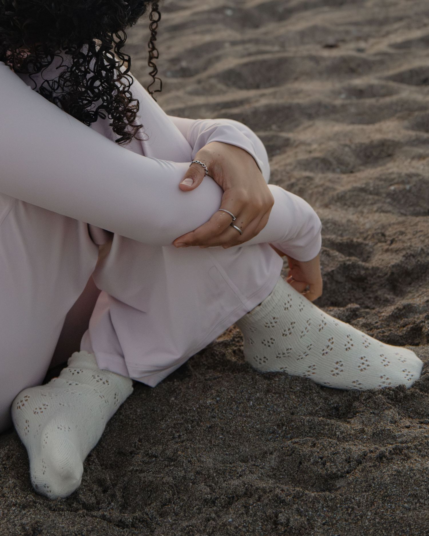 Person sitting on sand wearing light pink pants and white lace knit socks, with rings on fingers.