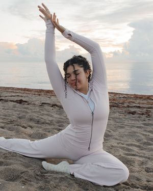 Woman in white athletic suit doing a yoga pose on a sandy beach at sunset with ocean in background.