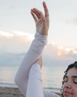 Woman in white long-sleeve top practicing yoga on beach at sunset with hand raised in mudra pose.