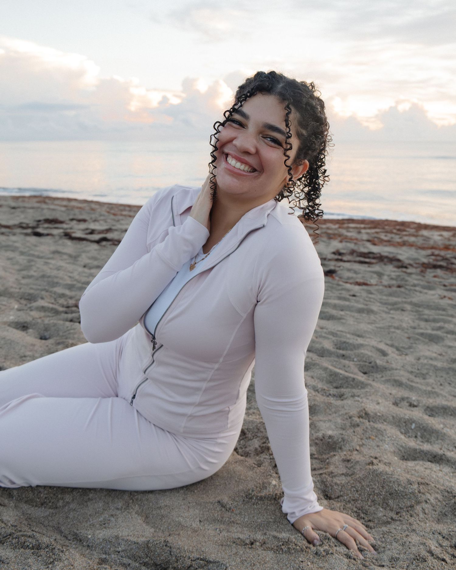 Smiling woman in white fitted jumpsuit sitting on sandy beach at sunset with curly hair.