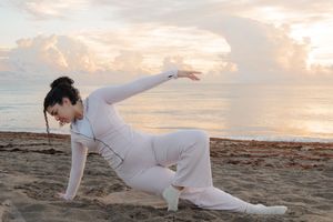 Woman in white outfit practicing yoga on a sandy beach at sunset with ocean and clouds in background.