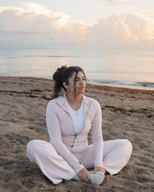 Young woman in white outfit sitting cross-legged on sandy beach at sunset with ocean waves in background.