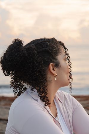 Woman with curly hair in ponytail gazes at sunset sky near ocean beach, wearing pink jacket and gold earring.