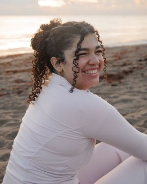 Smiling woman with curly hair in white top sitting on a sandy beach at sunset.