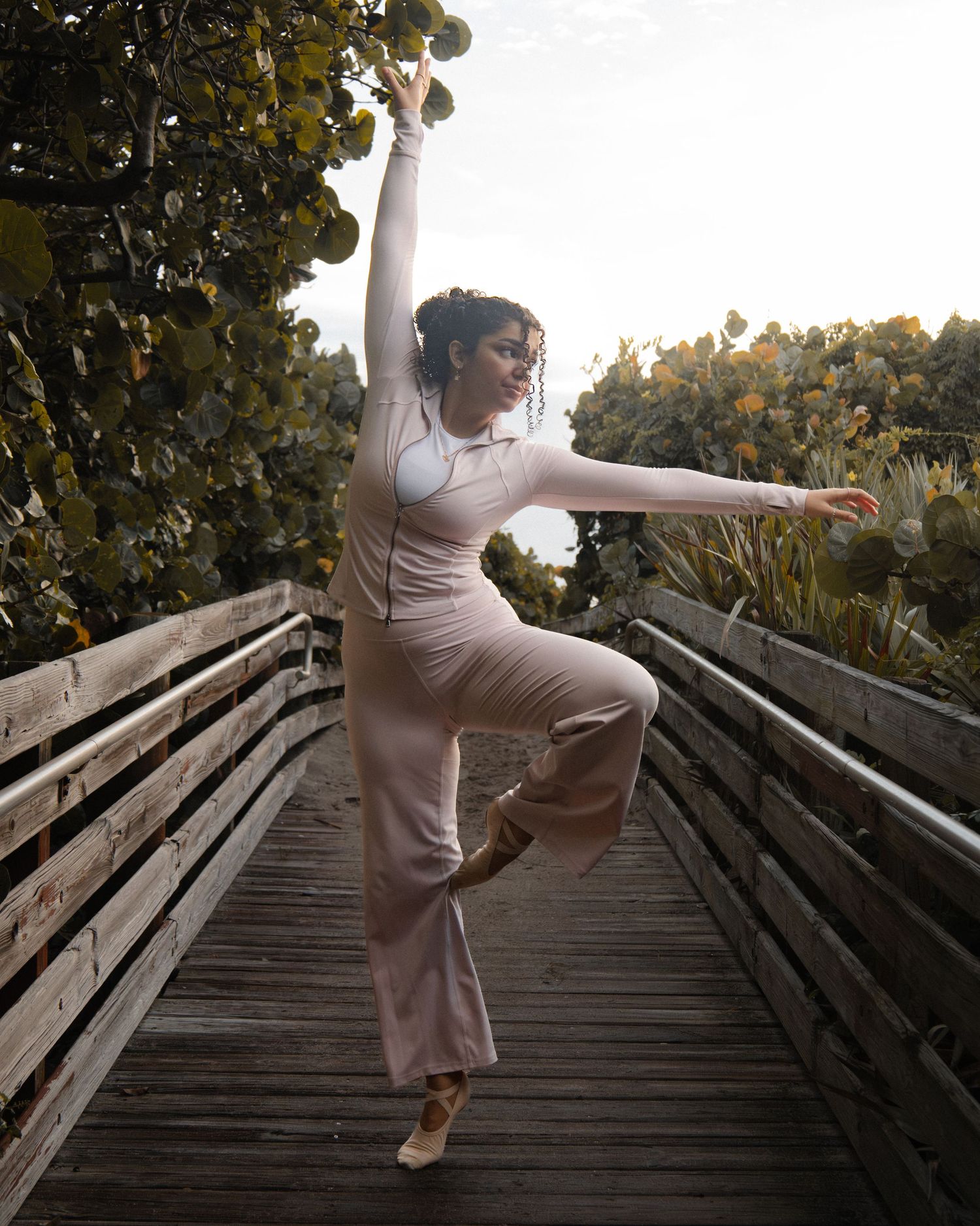 A dancer gracefully balances on one leg in a beige jumpsuit on a wooden bridge surrounded by lush green trees.