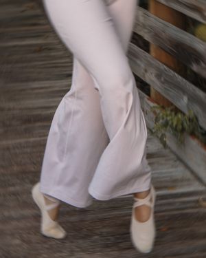 Blurred motion shot of a dancer in white wide-leg pants and ballet flats on a wooden bridge.
