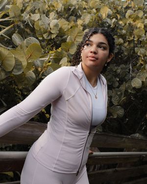 Woman in light pink athletic zip-up jacket poses outdoors with lush green foliage in the background.