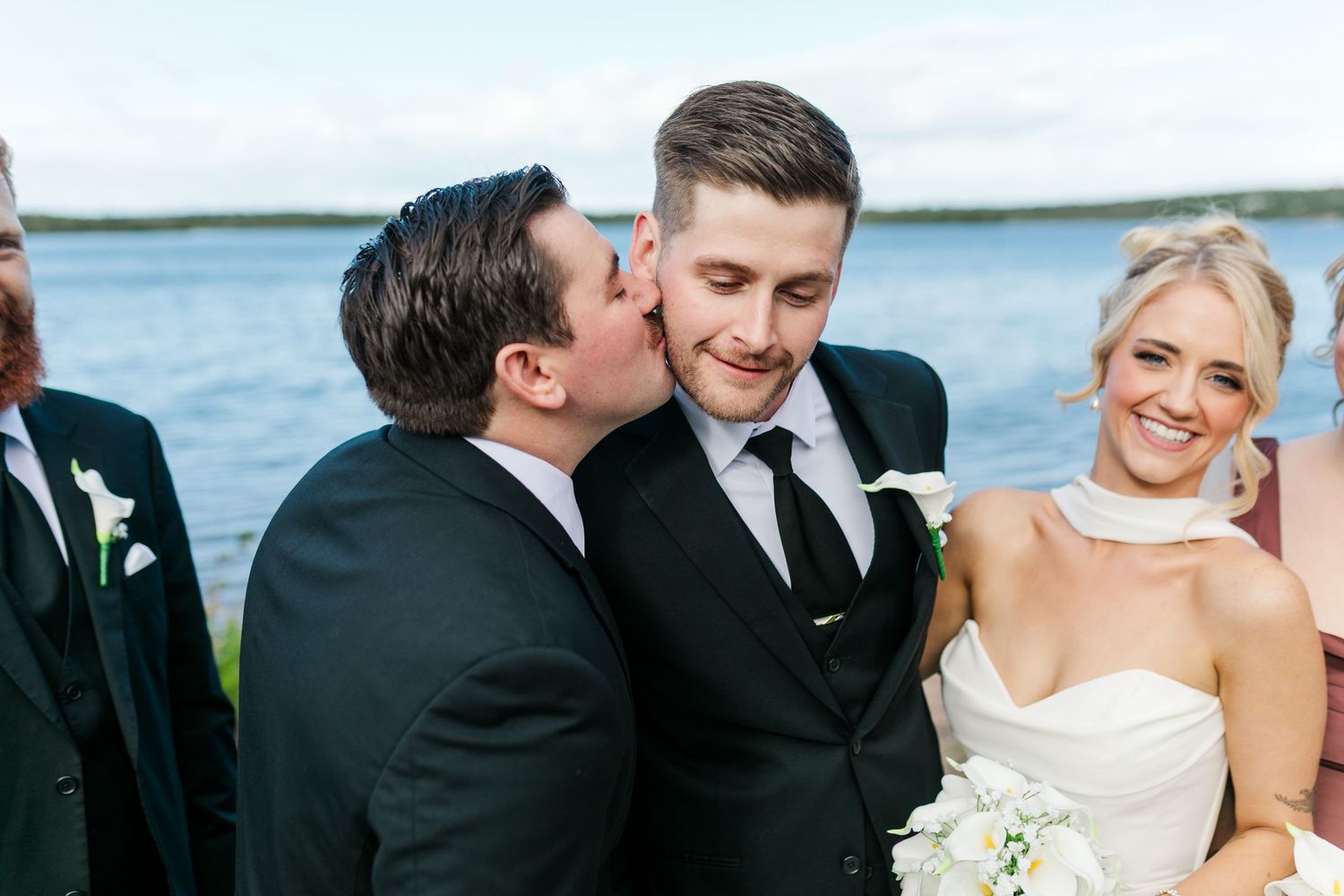 Groom receives a kiss on the cheek at outdoor waterfront wedding ceremony while smiling bride holds white bouquet.
