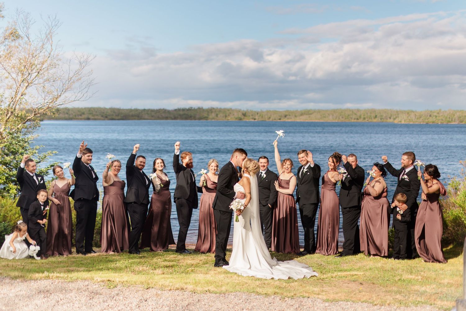 Large wedding party celebrating by a lake on a sunny day, bride and groom kissing at center.