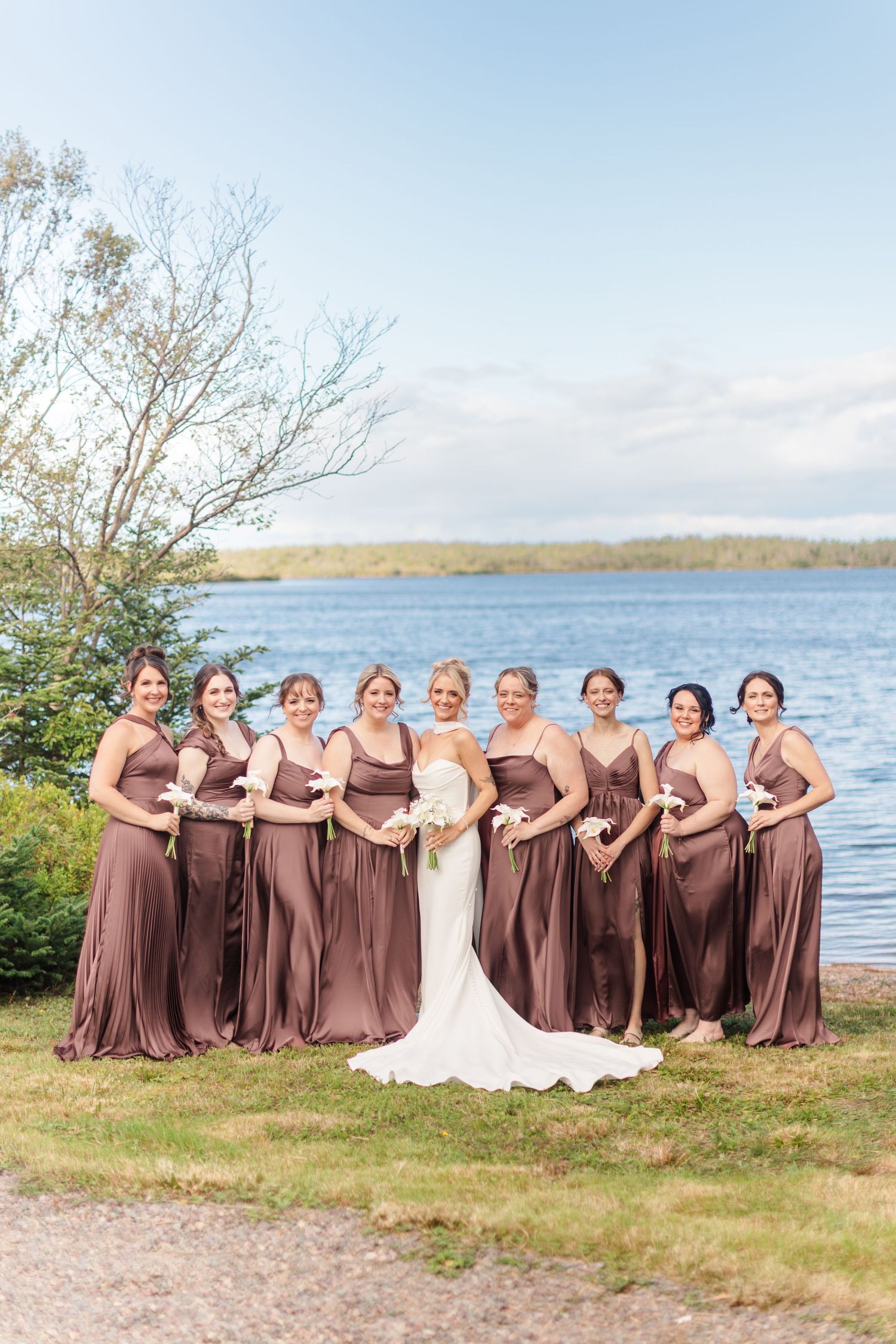Bride in white gown poses with nine bridesmaids in mauve satin dresses by a lakeside on a sunny day.