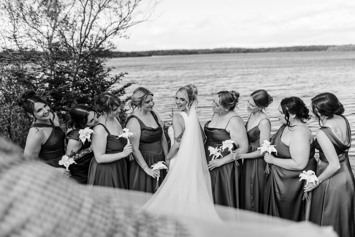 Black and white photo of bridesmaids in maxi dresses gathered around bride near a lake.