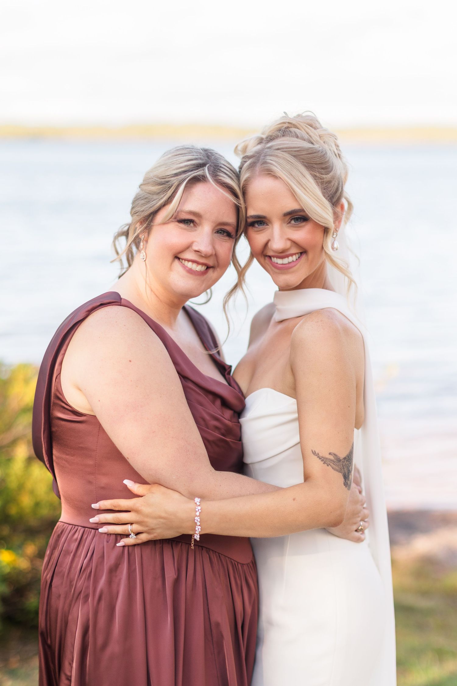 Bride in white gown embraces bridesmaid in mauve dress, smiling by a lakeside at an outdoor wedding.
