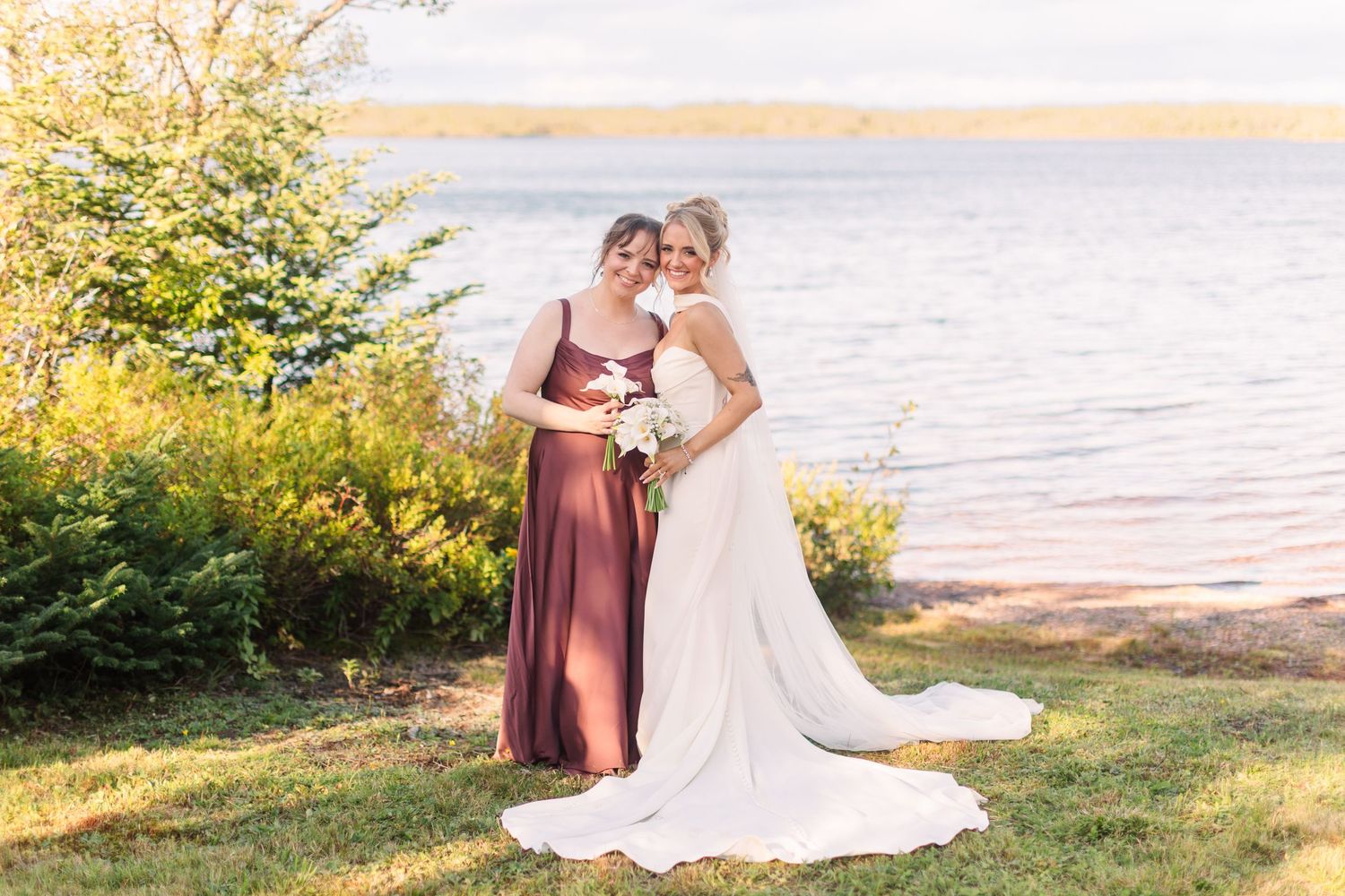 Bride and bridesmaid in burgundy gown posing by a lakeside, holding a bouquet on a sunny day.
