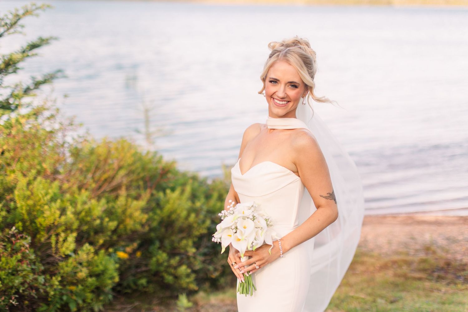 Smiling bride in strapless white gown holding bouquet by lakeside.