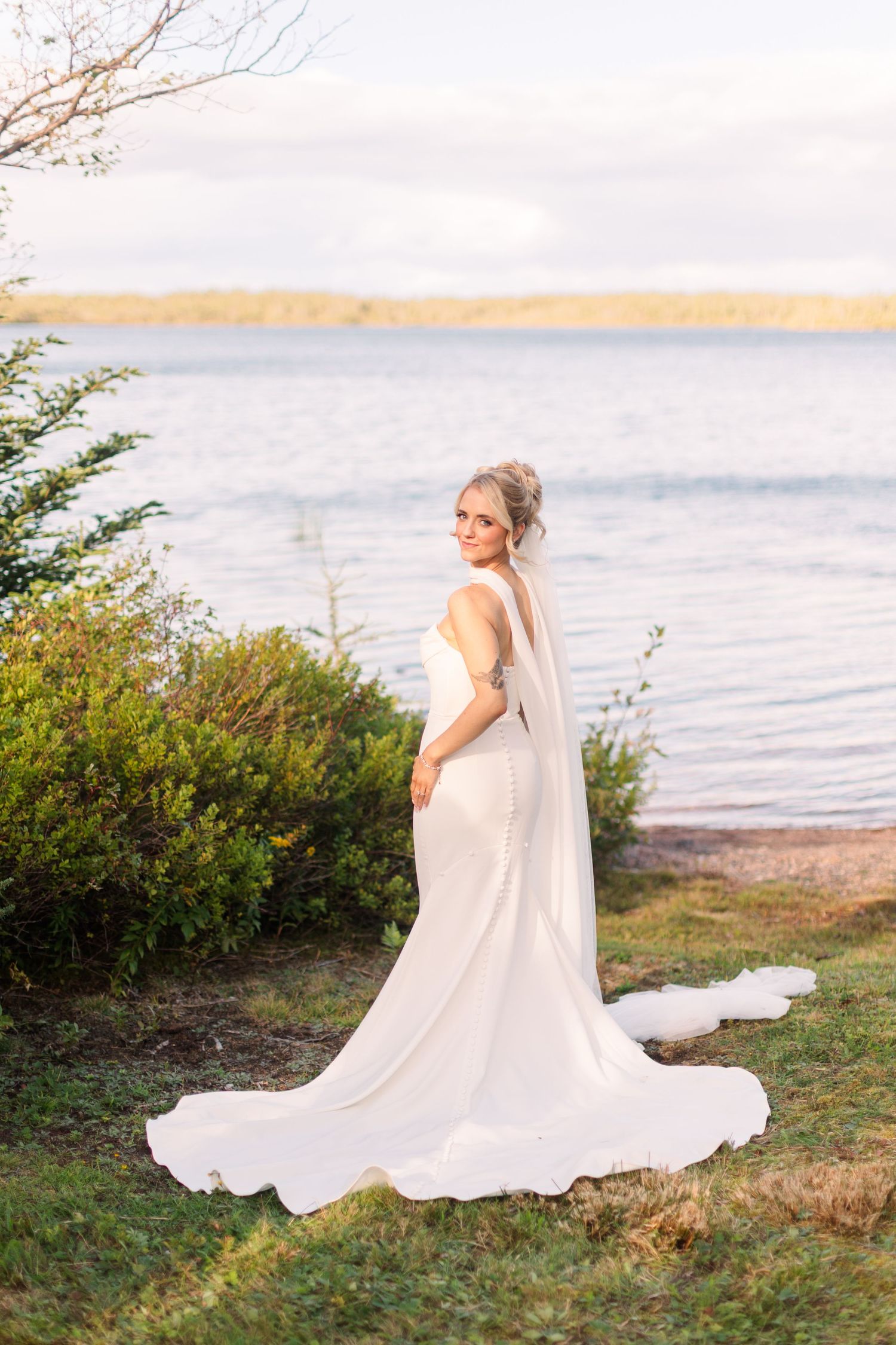 Bride in elegant white gown and veil poses by a serene lakeside, looking back over her shoulder.
