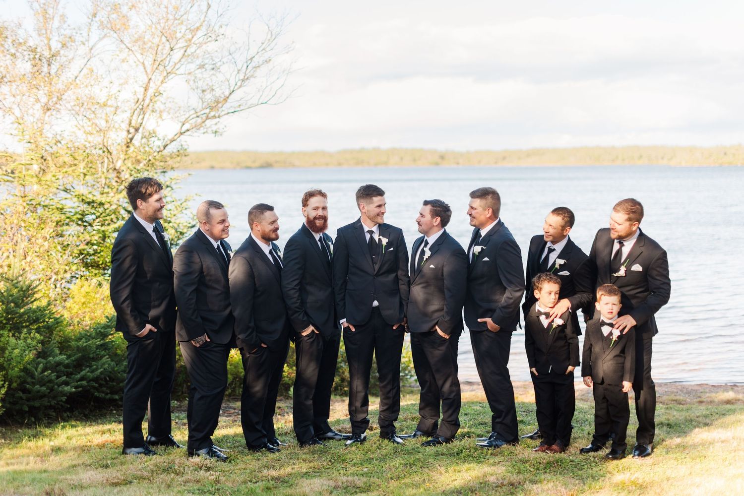 Groom and groomsmen in black suits stand together outdoors by a lake on a wedding day.