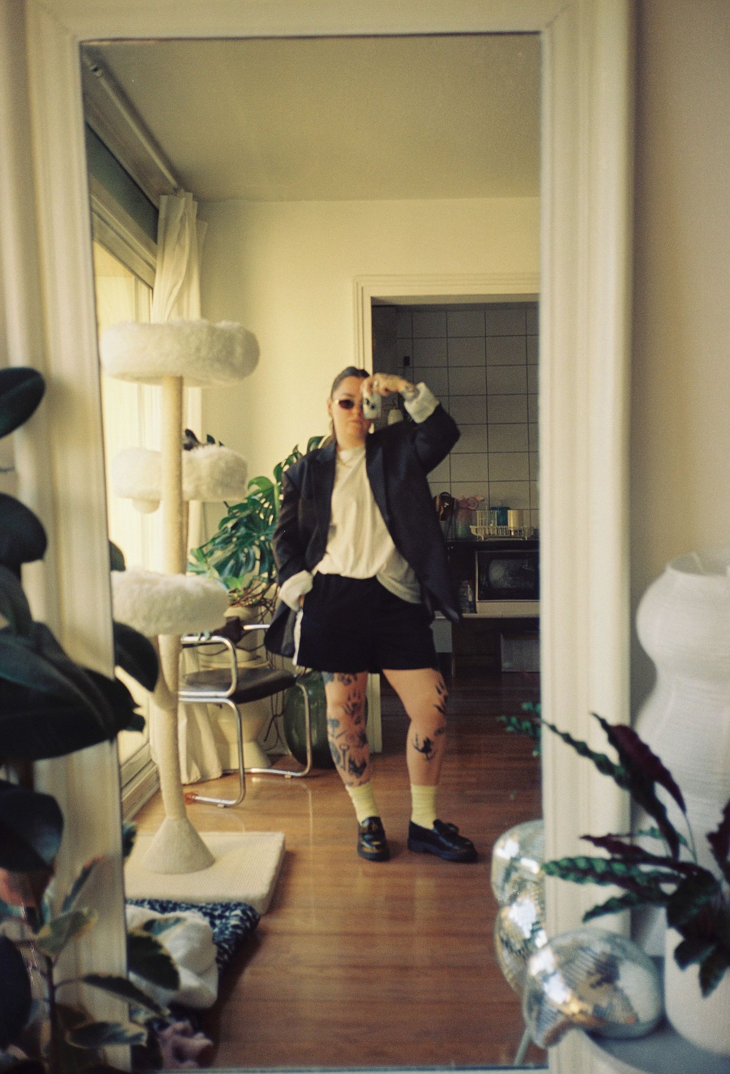 Person posing in doorway wearing black blazer, mini skirt, patterned tights, and loafers in a cozy apartment.