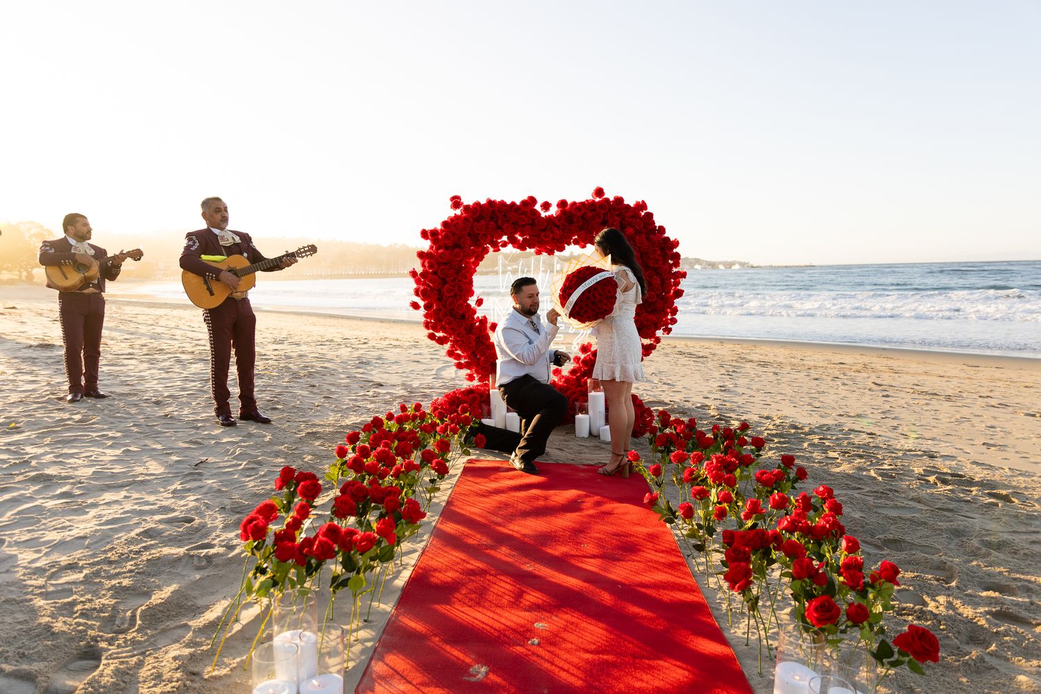 Man proposes to woman on a romantic red rose-lined beach setup with mariachi band at sunset.