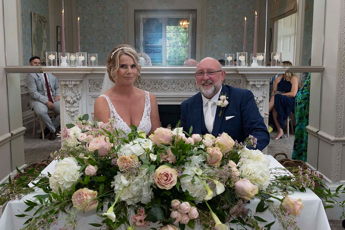 Bride and groom seated behind a display of cream and pink flowers during their wedding ceremony at the Belsfield Hotel.