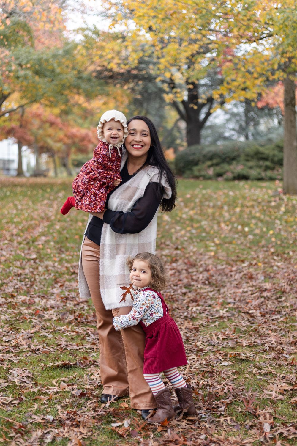 A smiling mother holds her baby girl while her toddler daughter hugs her leg in a colorful autumn park.
