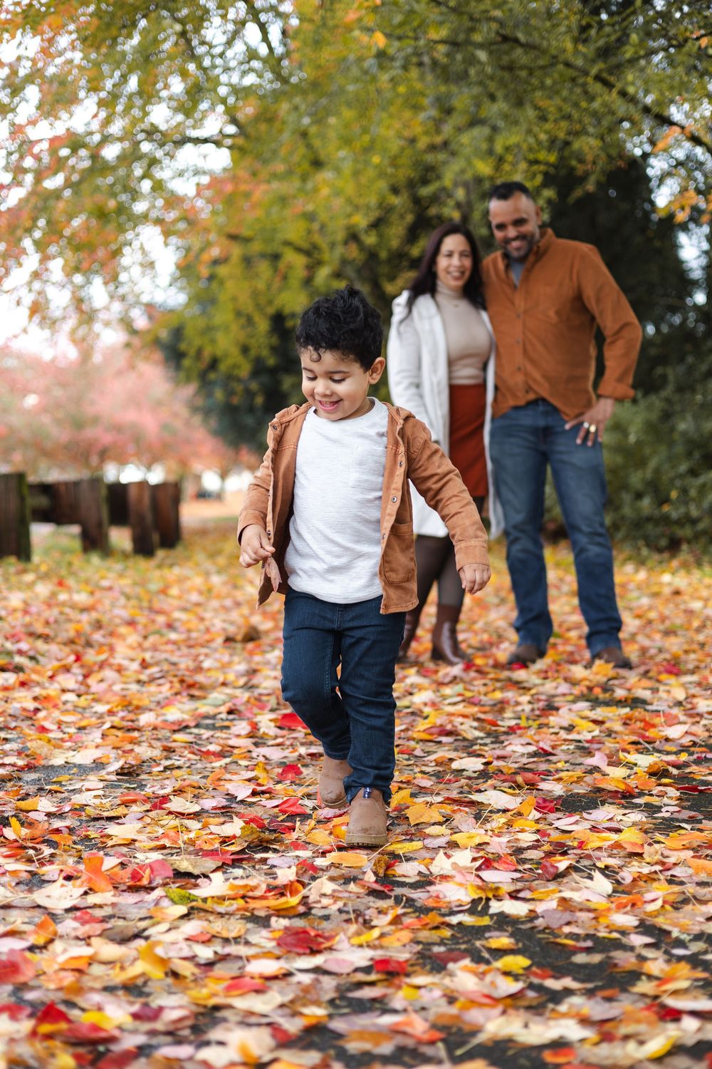 A joyful boy runs through a colorful autumn park while his smiling parents watch fondly in the background.