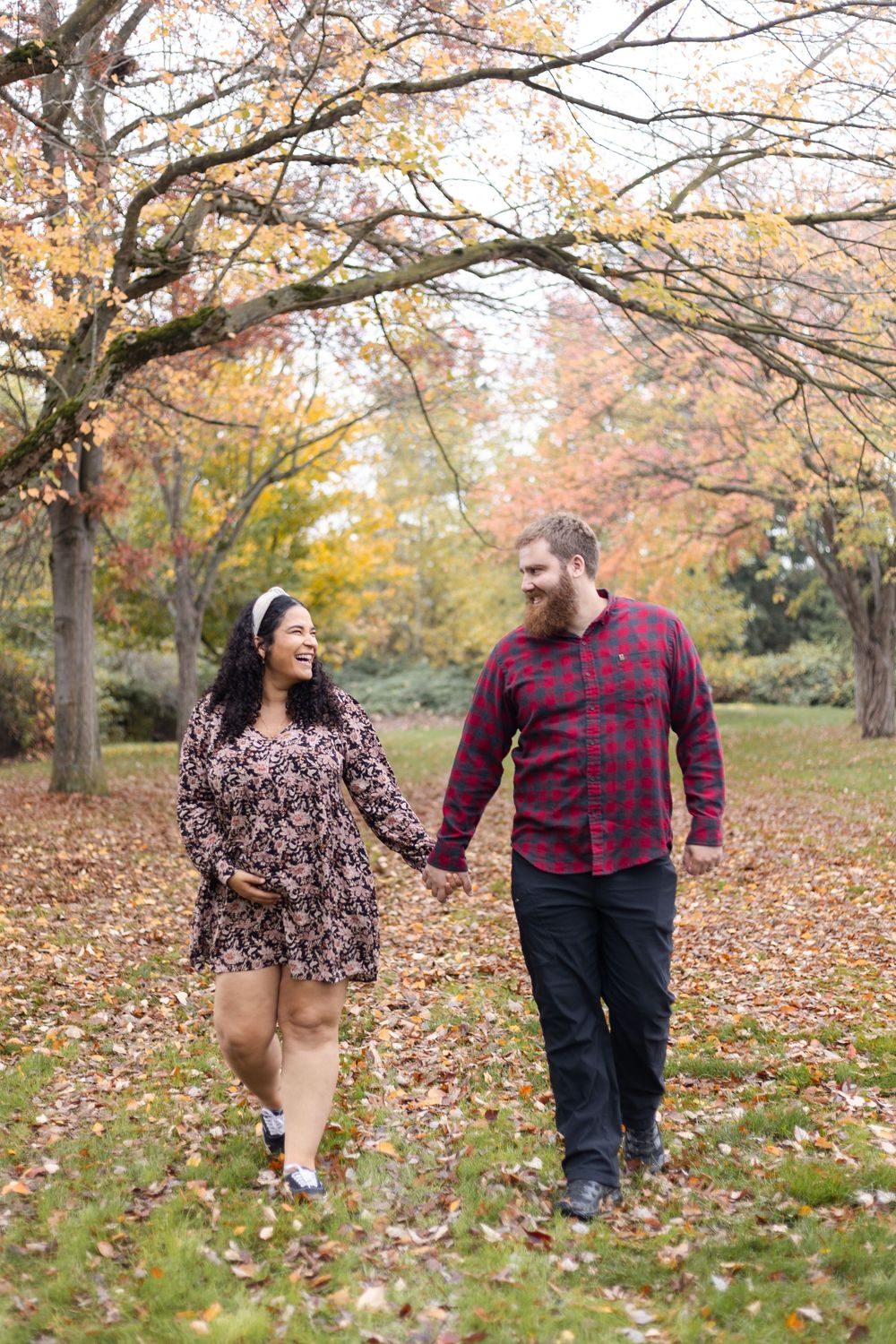 Couple holding hands and smiling while walking through a park with colorful autumn foliage.