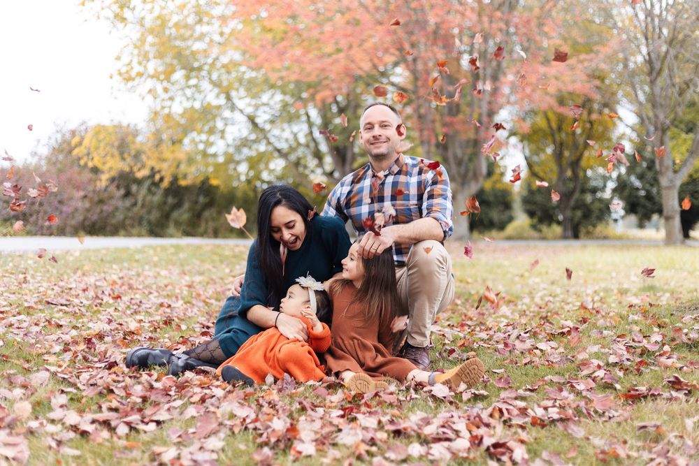 Family sitting on fall leaves in a park, laughing as leaves fall around them on a sunny autumn day.