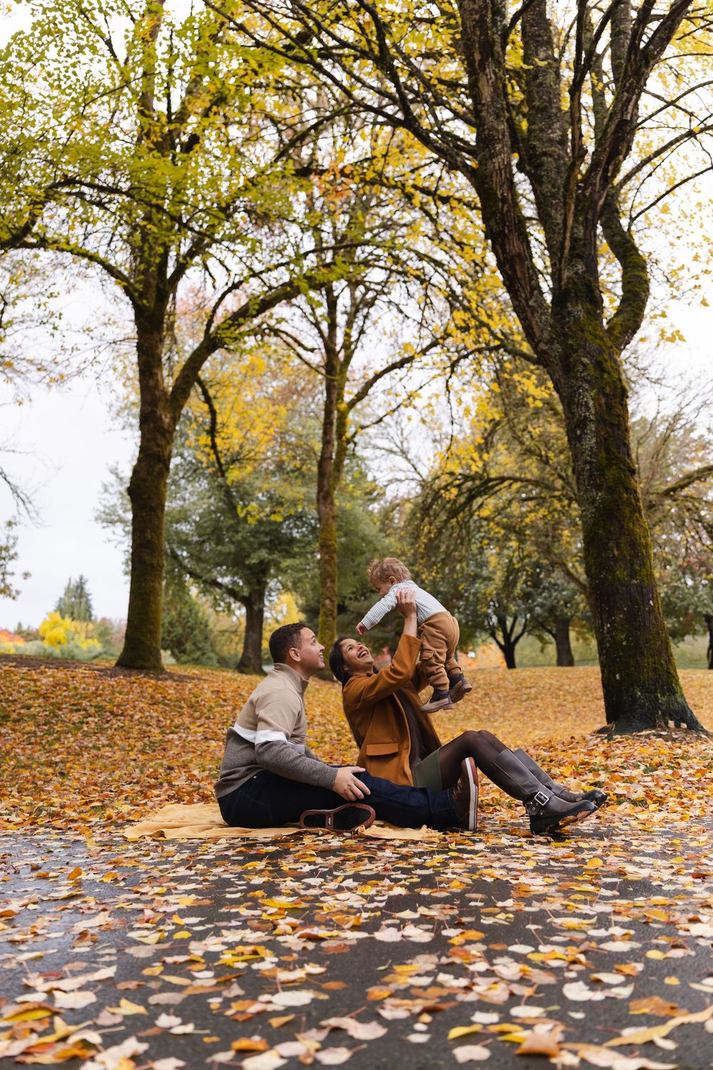 Family sitting on fallen autumn leaves in a park, parents lifting their baby up while surrounded by golden trees.