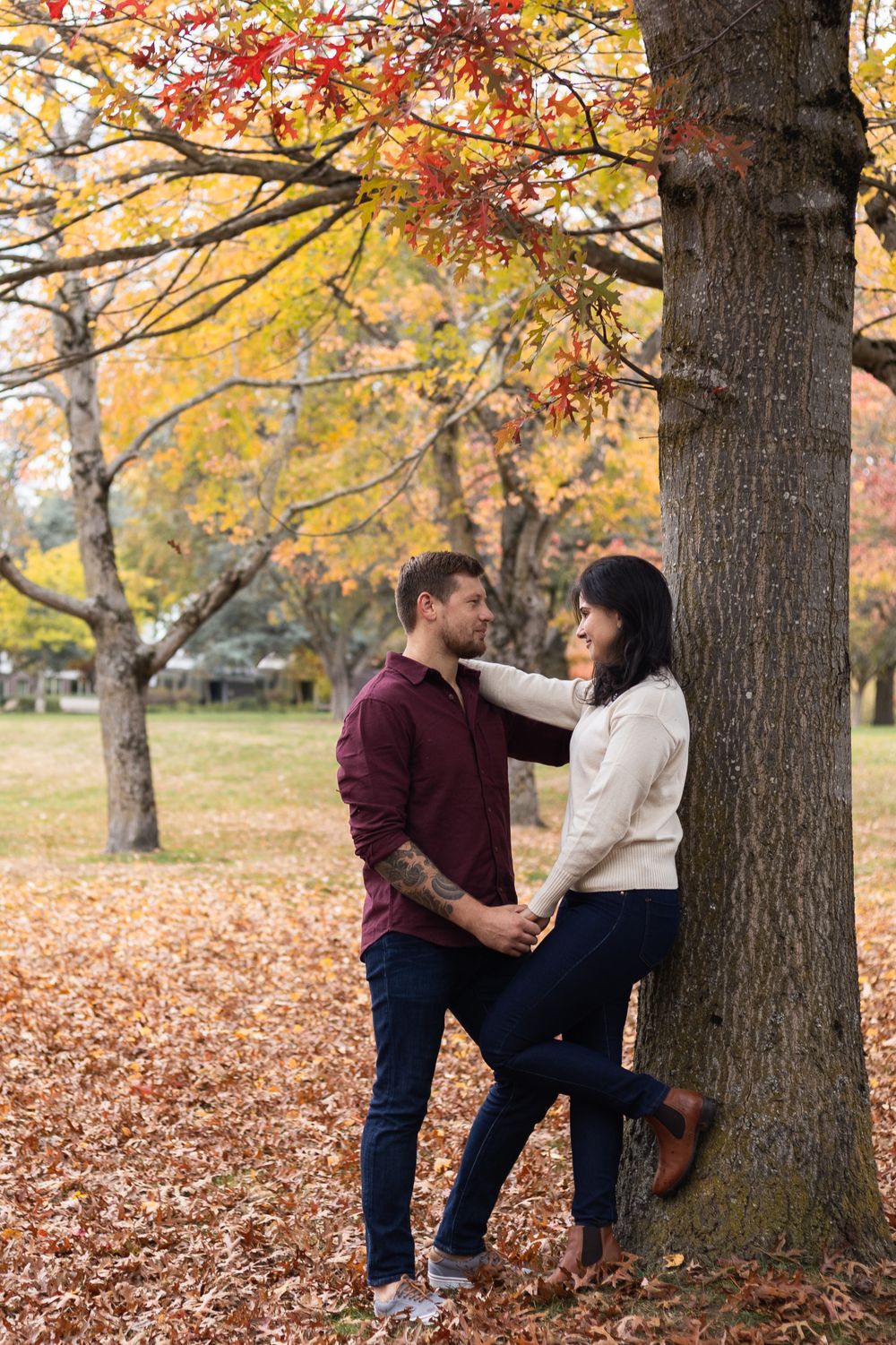 Couple holding hands and gazing at each other leaning against a tree in a park during autumn.