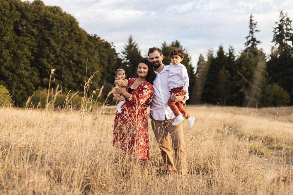 Family of four posing in golden field with evergreen trees, parents holding two young children outdoors.