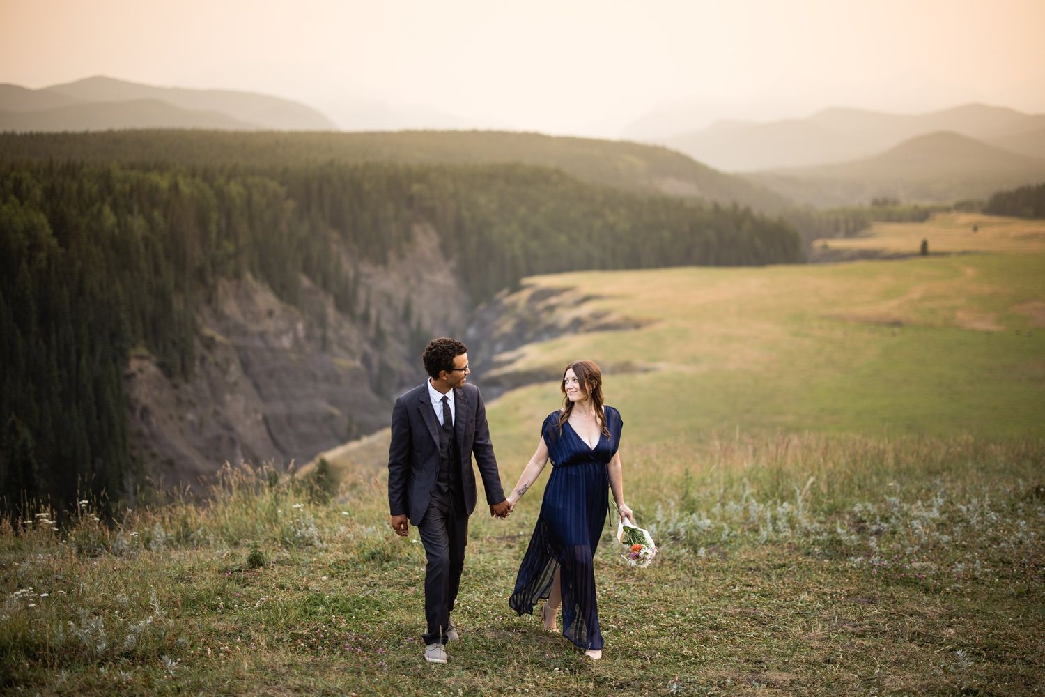 Engaged couple walking hand in hand through a mountain valley meadow with forested hills behind