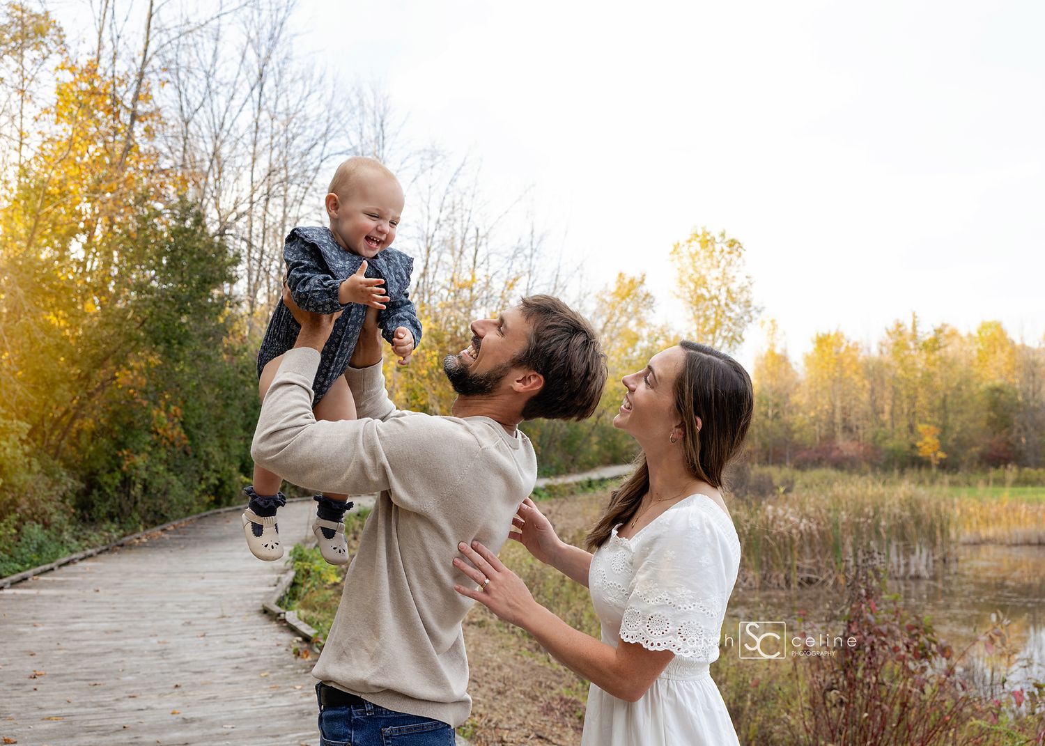 A happy young family with a bearded father lifting a laughing baby outdoors in an autumn park setting.