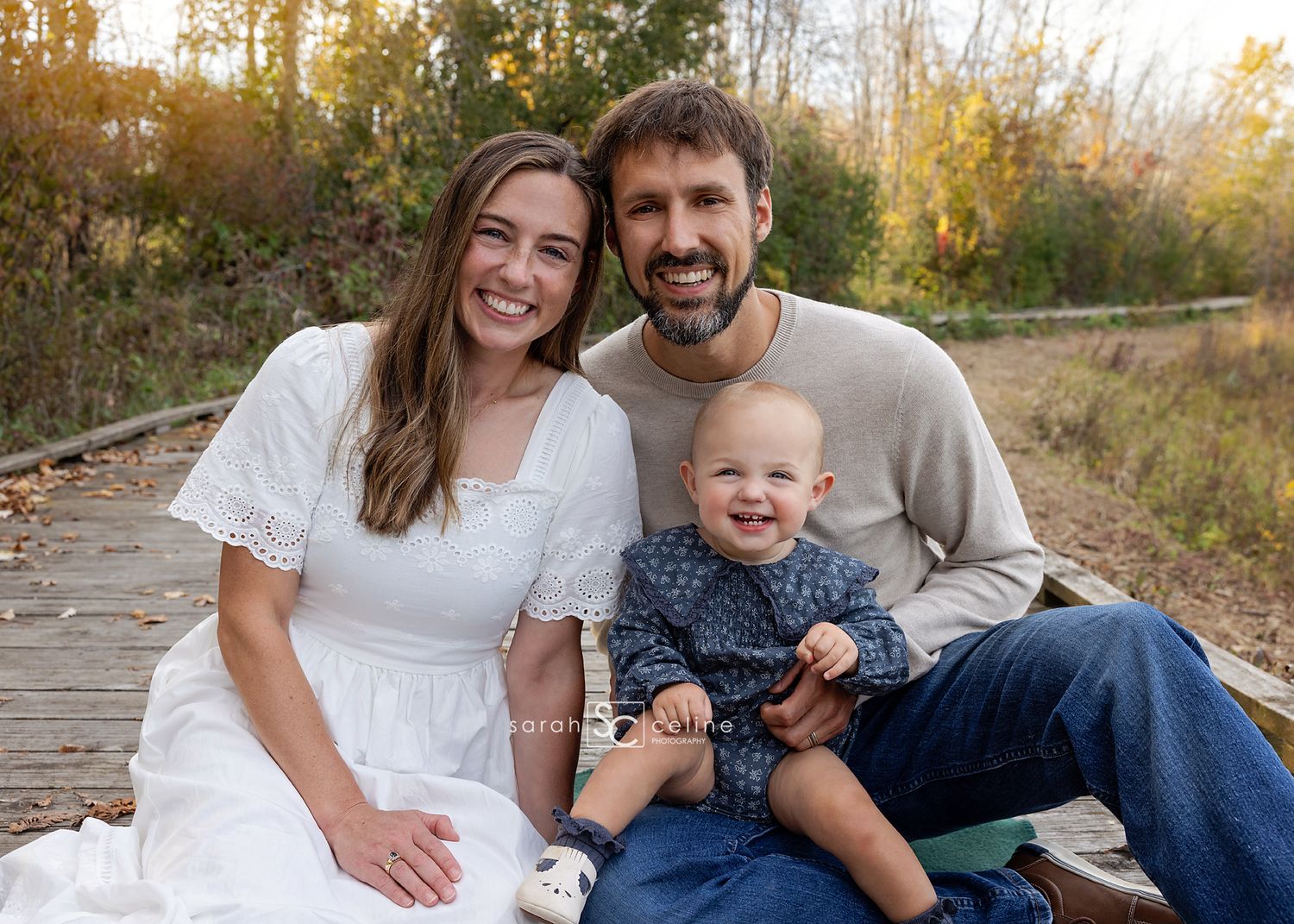 Happy family of three posing outdoors on a wooden path, parents smiling with baby in fall setting.