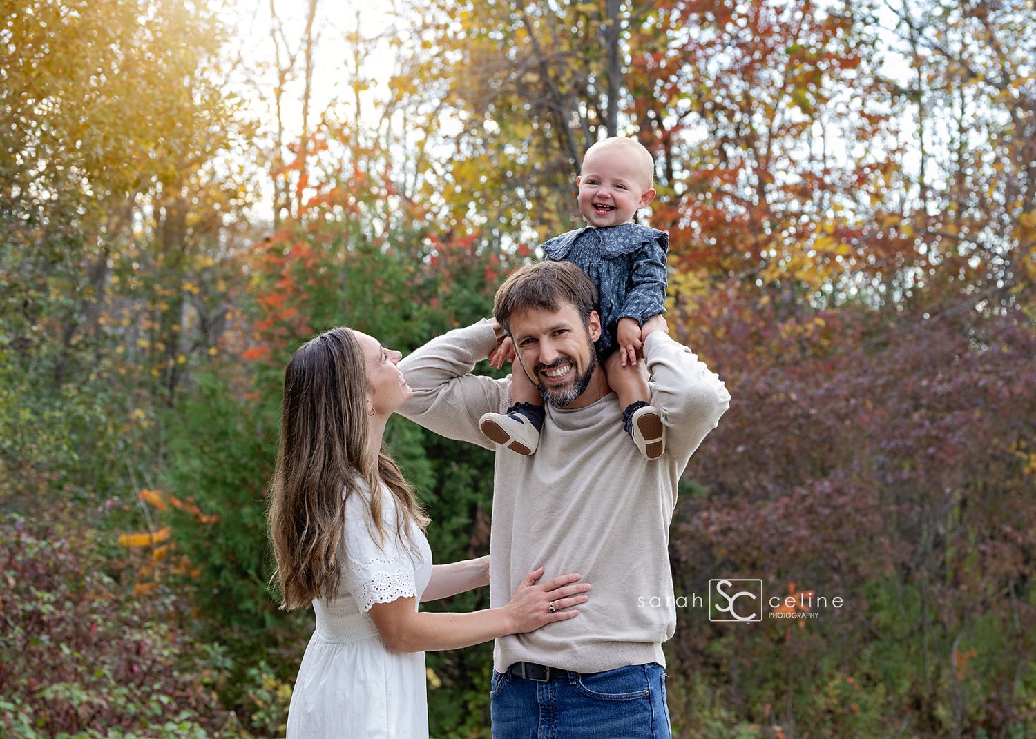 Family portrait with baby on dad's shoulders, mom in white dress, autumn foliage background.