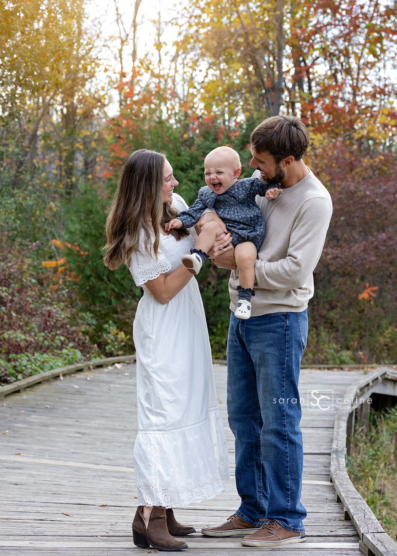Family of three on a wooden bridge in autumn, parents smiling at laughing baby in navy dress.