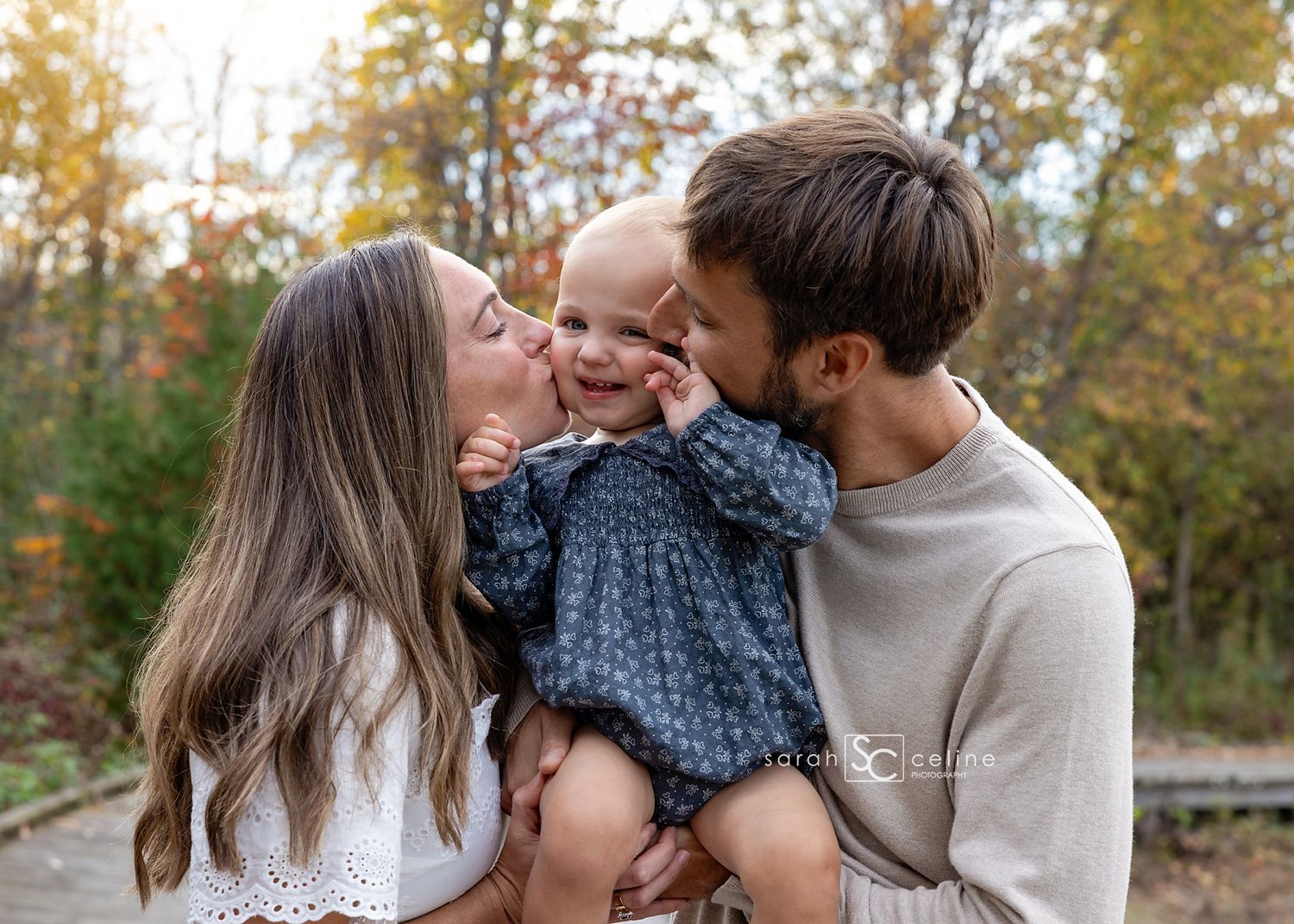 Parents kissing their smiling baby girl on cheeks outdoors in autumn setting with colorful fall foliage background.