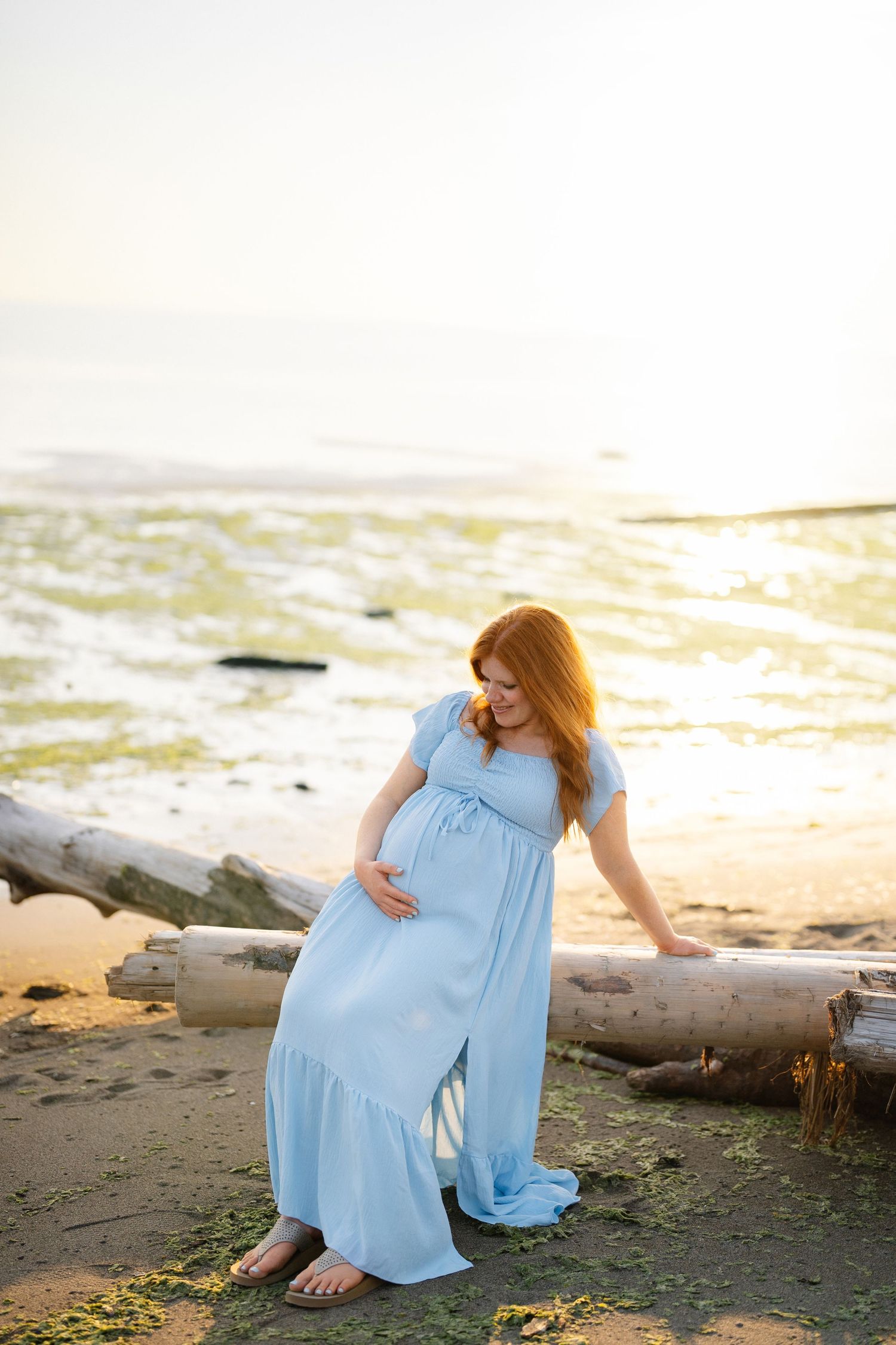 Pregnant woman in a flowing light blue maternity dress sits on a driftwood log on the beach at sunset, resting a hand on her belly.