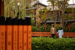 Couple holding hands on a Japanese garden path with traditional wooden buildings, red lanterns, green shrubs and orange inscribed posts.