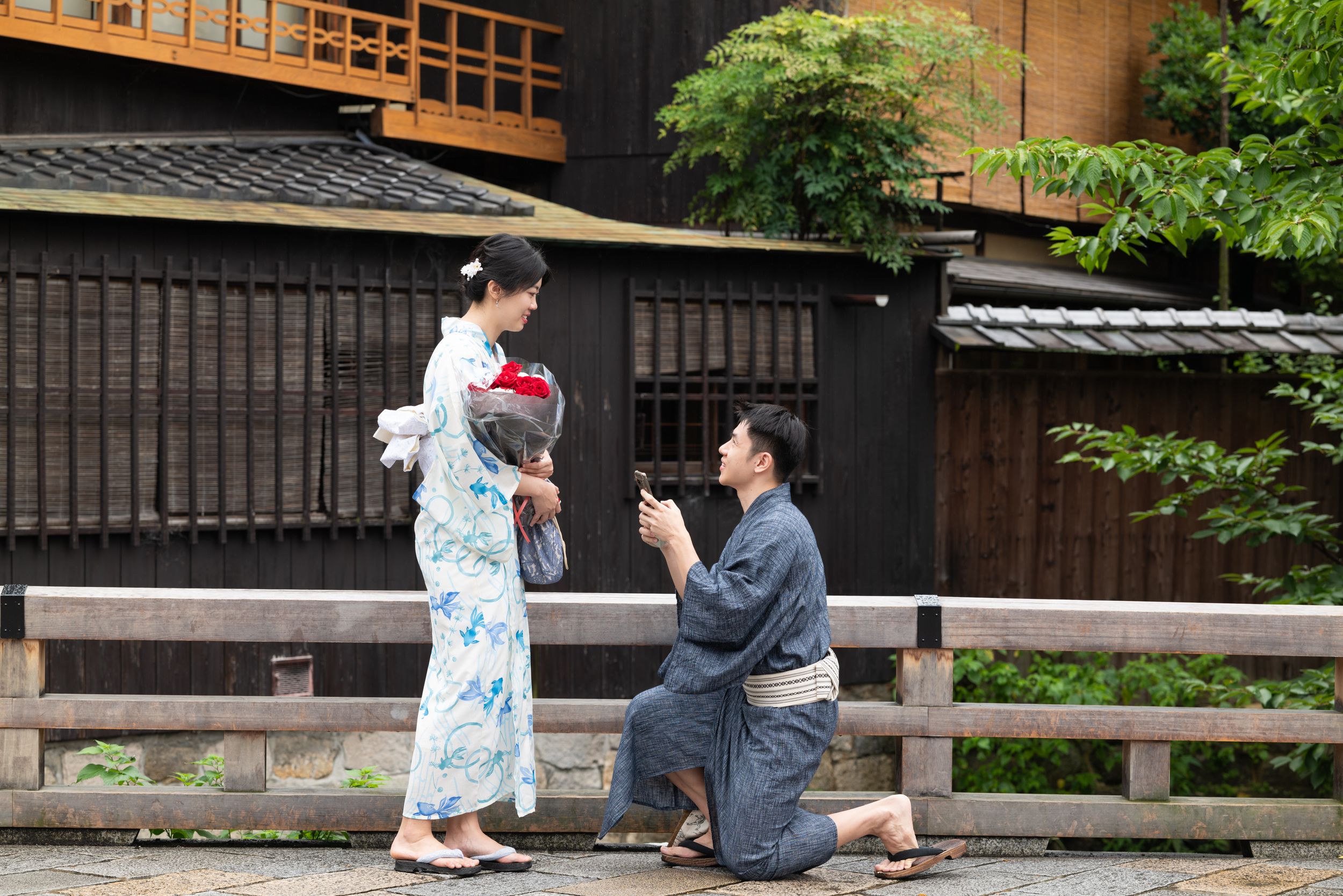 Man in a traditional Japanese kimono kneels on one knee proposing to a woman in a floral yukata holding a bouquet outside a wooden building.