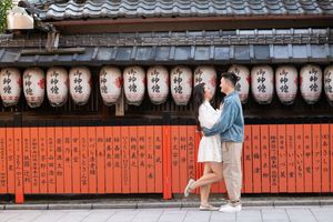 Couple embracing and kissing in front of a vermilion wooden fence lined with white paper lanterns on a traditional street in Kyoto, Japan.