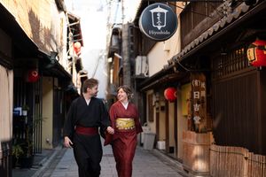 Couple in traditional Japanese kimono walking hand in hand down a quiet Kyoto lane lined with wooden buildings and red paper lanterns.
