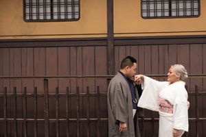 Couple in traditional Japanese kimono embracing in front of a rustic wooden fence and wall, the woman in white with a pink obi.