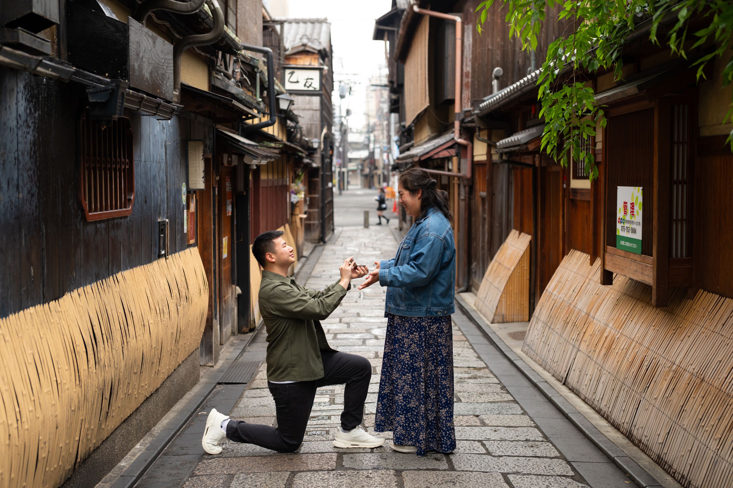Man kneeling on one knee proposes to a woman in a denim jacket on a narrow traditional Kyoto street lined with wooden buildings.