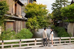 Couple stands on a wooden bridge over a calm canal, surrounded by lush green trees and traditional Japanese wooden houses in Kyoto.